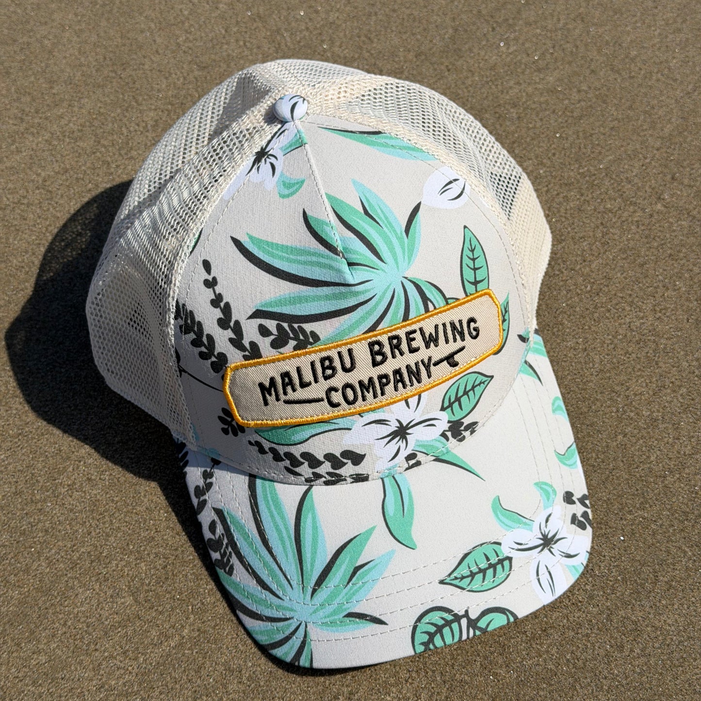 Trucker hat with tropical leaf pattern and Malibu Brewing Company logo on sand