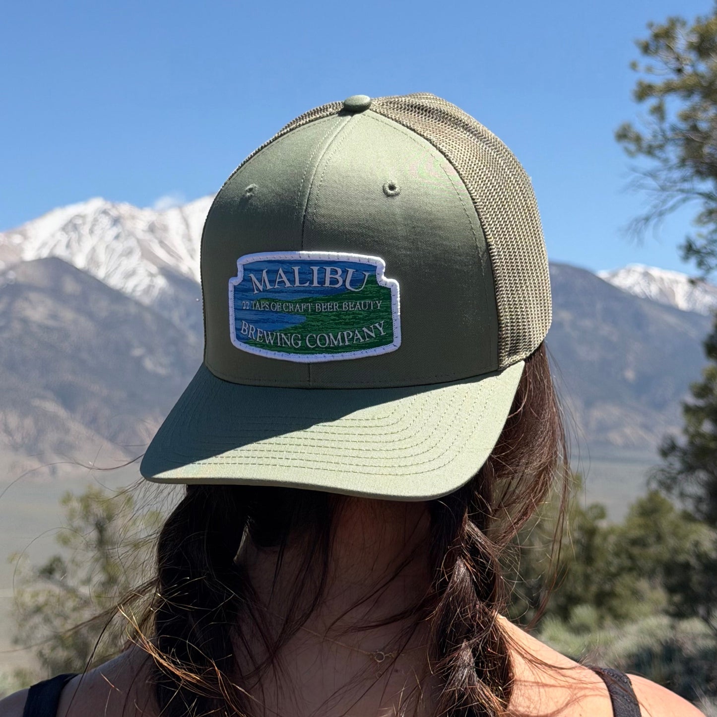 A person with brown braided hair wears an olive green Malibu City Sign Hat with an embroidered patch, standing outdoors against snow-capped mountains, trees, and a clear blue sky.