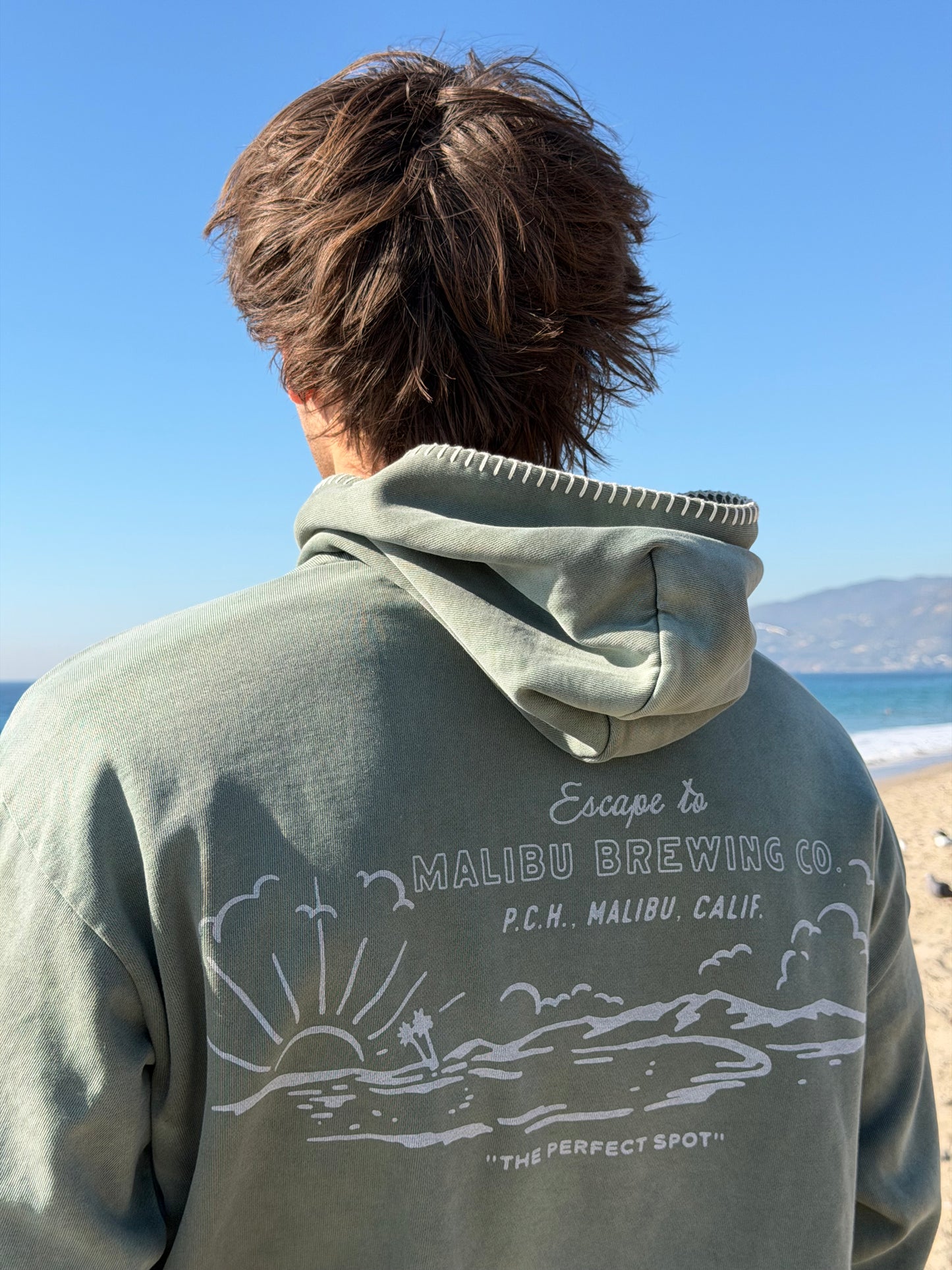 Person wearing a hoodie with 'Escape Malibu Brewing Co.' text and graphics against a clear blue sky.