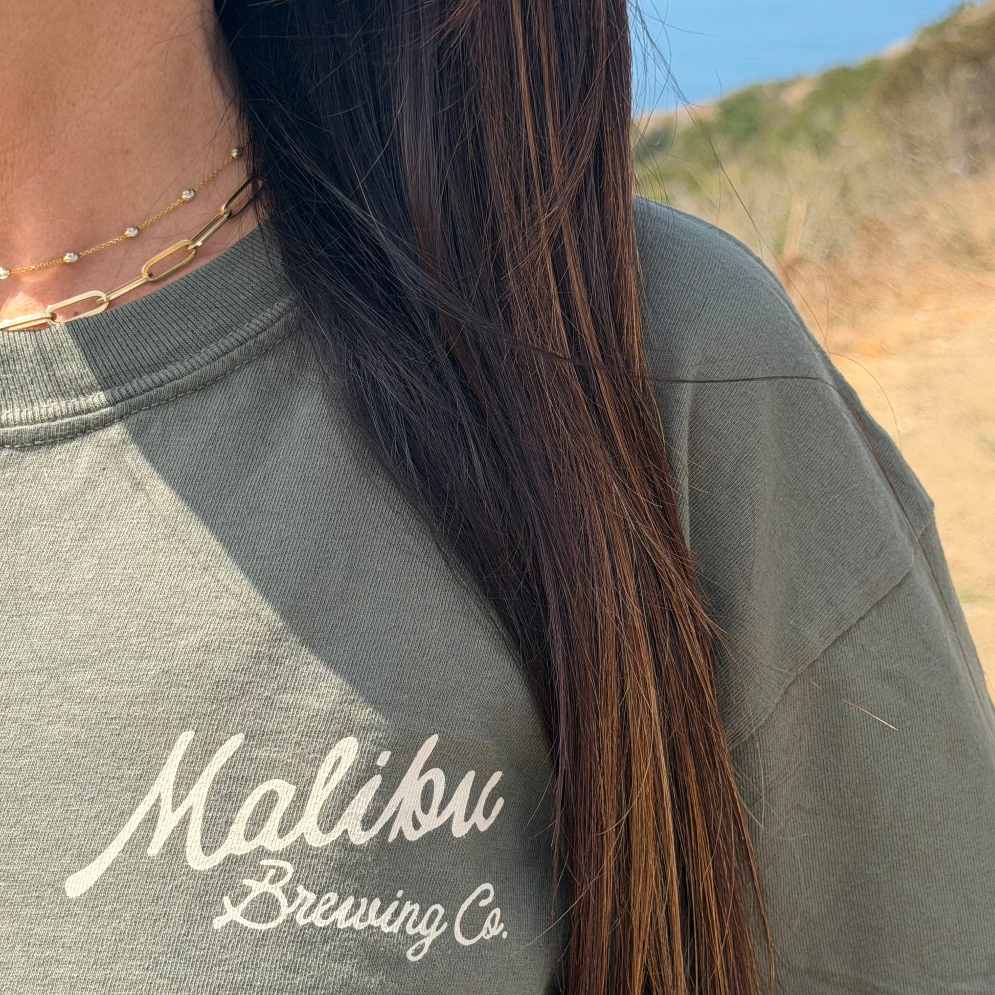 Close-up of a person in the green Crafted in Paradise Tee and layered gold necklaces, long brown hair draped over their shoulder. This unisex coastal shirt pops against a softly blurred outdoor backdrop of greenery and sky.