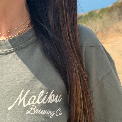 Close-up of a person in the green Crafted in Paradise Tee and layered gold necklaces, long brown hair draped over their shoulder. This unisex coastal shirt pops against a softly blurred outdoor backdrop of greenery and sky.
