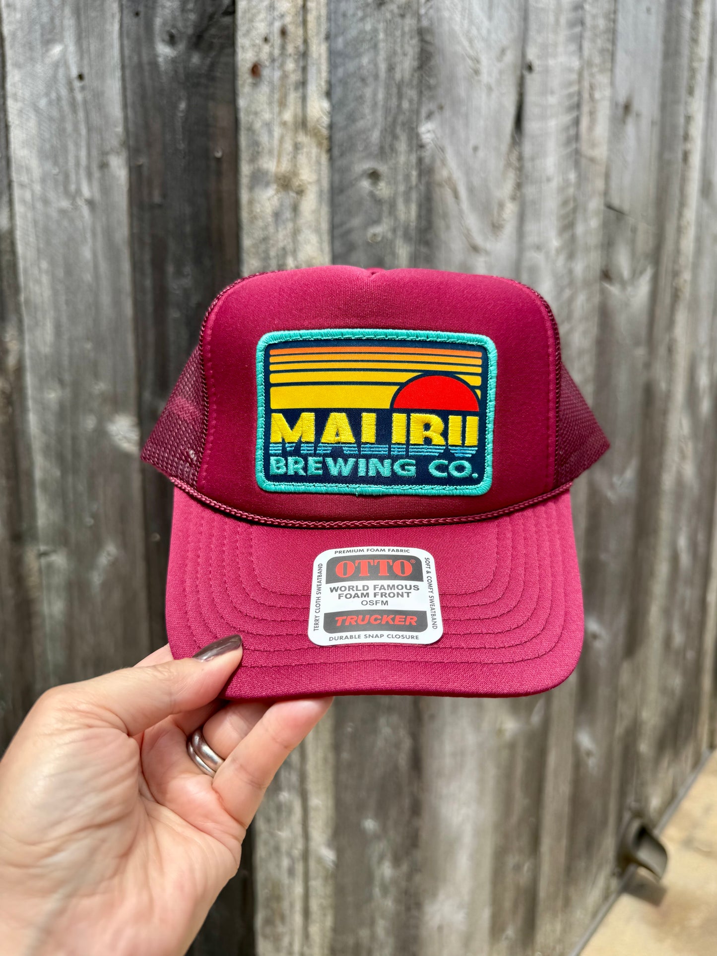 A hand holds the Otto Sunset Patch Hat, a maroon trucker hat with a bold Malibu Brewing Co. patch on the front and an OTTO brand sticker on the bill, set against a wooden fence backdrop—perfect for summer style.