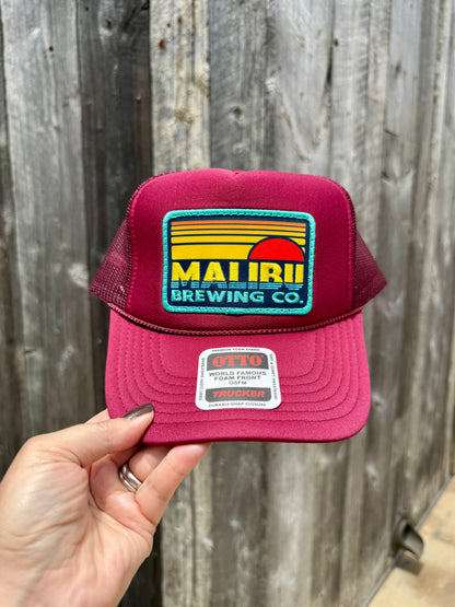 A hand holds the Otto Sunset Patch Hat, a maroon trucker hat with a bold Malibu Brewing Co. patch on the front and an OTTO brand sticker on the bill, set against a wooden fence backdrop—perfect for summer style.