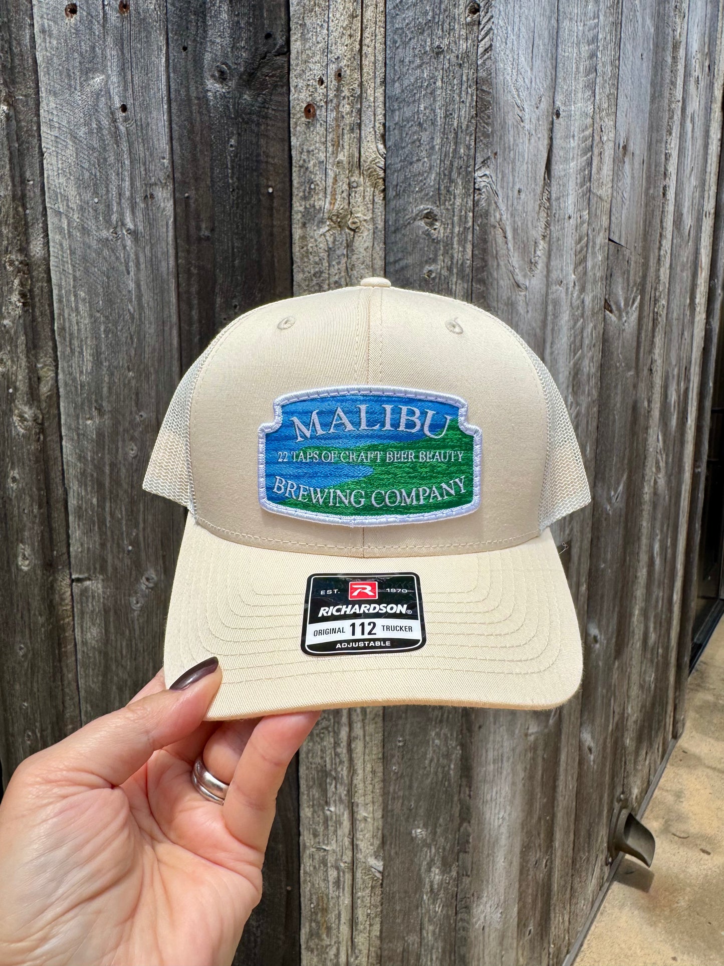 A hand holds a beige Malibu City Sign Hat with an embroidered patch reading "Malibu Brewing Company—27 Taps of Craft Beer Beauty," featuring a black Richardson 112 sticker on the brim, set against a wooden wall background.