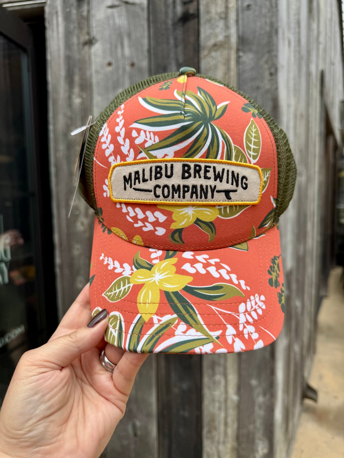 A hand holds the Floral Trucker Hat, featuring coral color, floral leaf patterns, an adjustable back, and a “Malibu Brewing Company” patch, shown against a weathered wooden wall.
