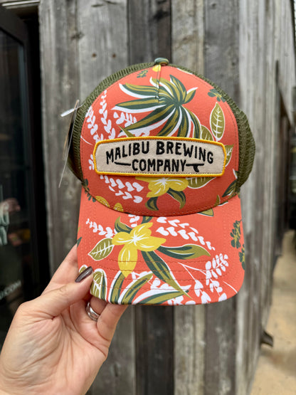 A hand holds the Floral Trucker Hat, featuring coral color, floral leaf patterns, an adjustable back, and a “Malibu Brewing Company” patch, shown against a weathered wooden wall.