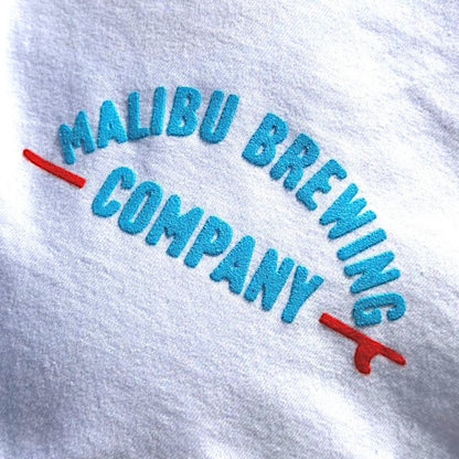 The Red, White & 'Bu Tee is a white unisex shirt featuring MALIBU BREWING COMPANY embroidered in blue, with "COMPANY" underlined in red ending in a curved tail—your perfect go-to summer tee.