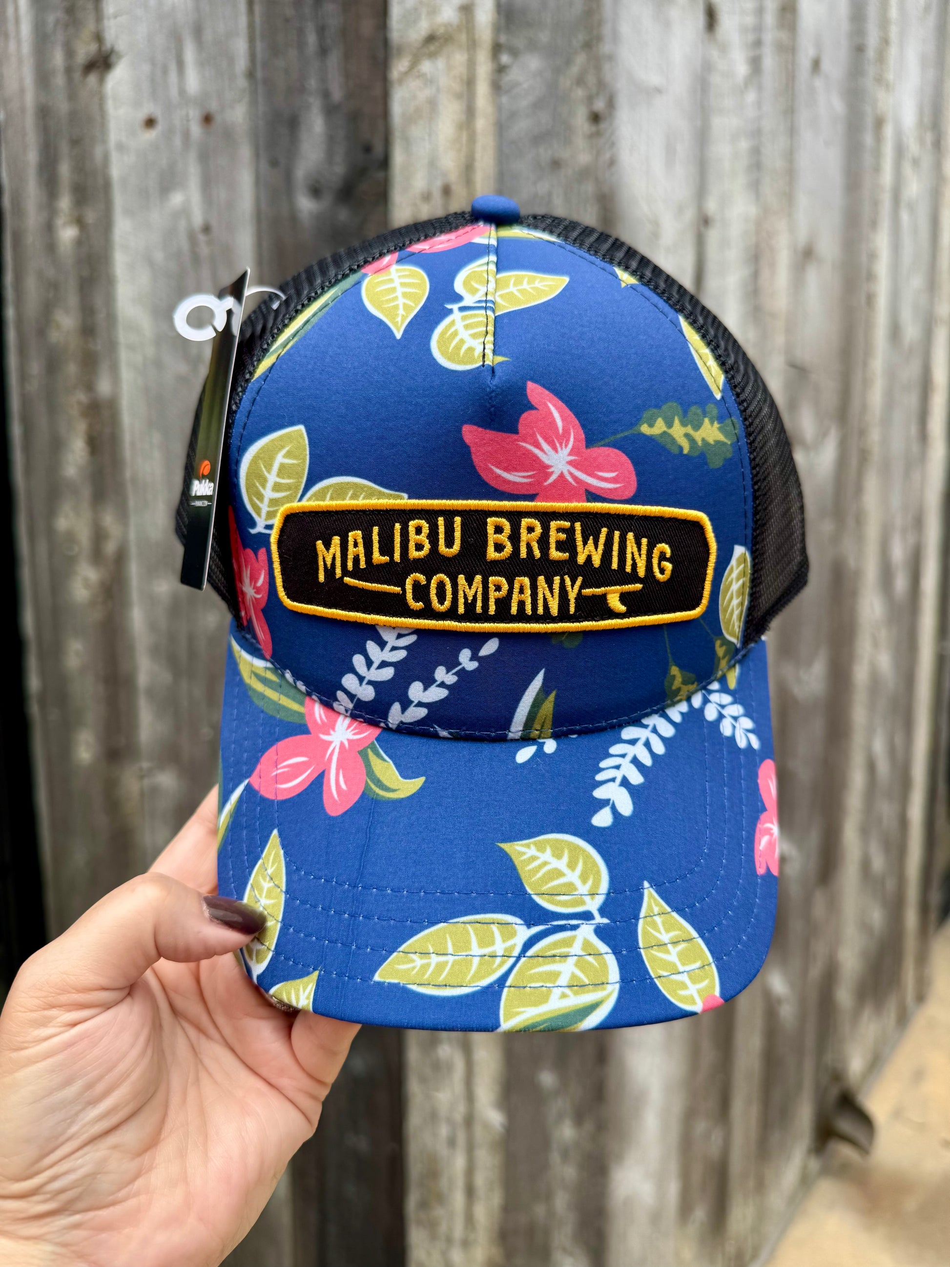 A hand holds the Floral Trucker Hat, featuring a blue floral design and an embroidered Malibu Brewing Company patch, against a background of weathered wood.