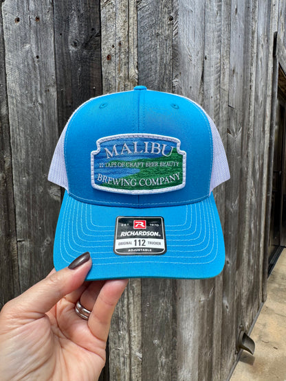 A hand holds a blue and white Malibu City Sign Hat with an embroidered Malibu Brewing Company patch, set against a rustic wooden wall. The Richardson snap-back cap displays the classic 112 Trucker sticker on its brim.