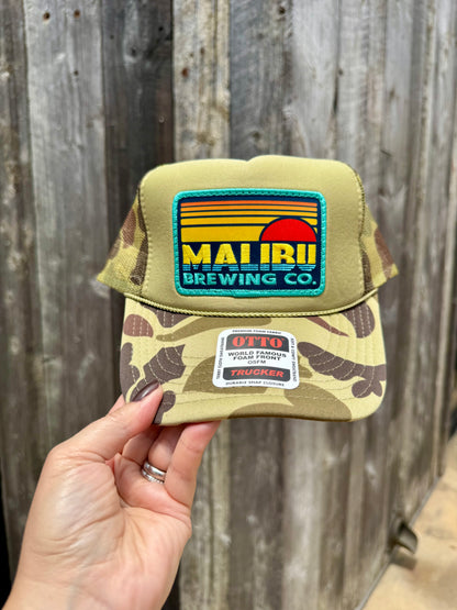 A hand holds the Otto Sunset Patch Hat, a camo trucker cap featuring a vibrant Mairii Brewing Co. patch and OTTO Trucker label, in front of a wooden fence—an ideal choice for your summer wardrobe.
