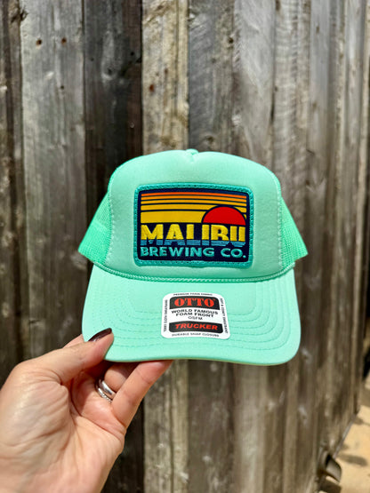 A hand holds the Otto Sunset Patch Hat, a mint green hat with retro stripes and a sun patch reading "Malibu Brewing Co."; a white tag is on the brim with a wooden fence in the background.
