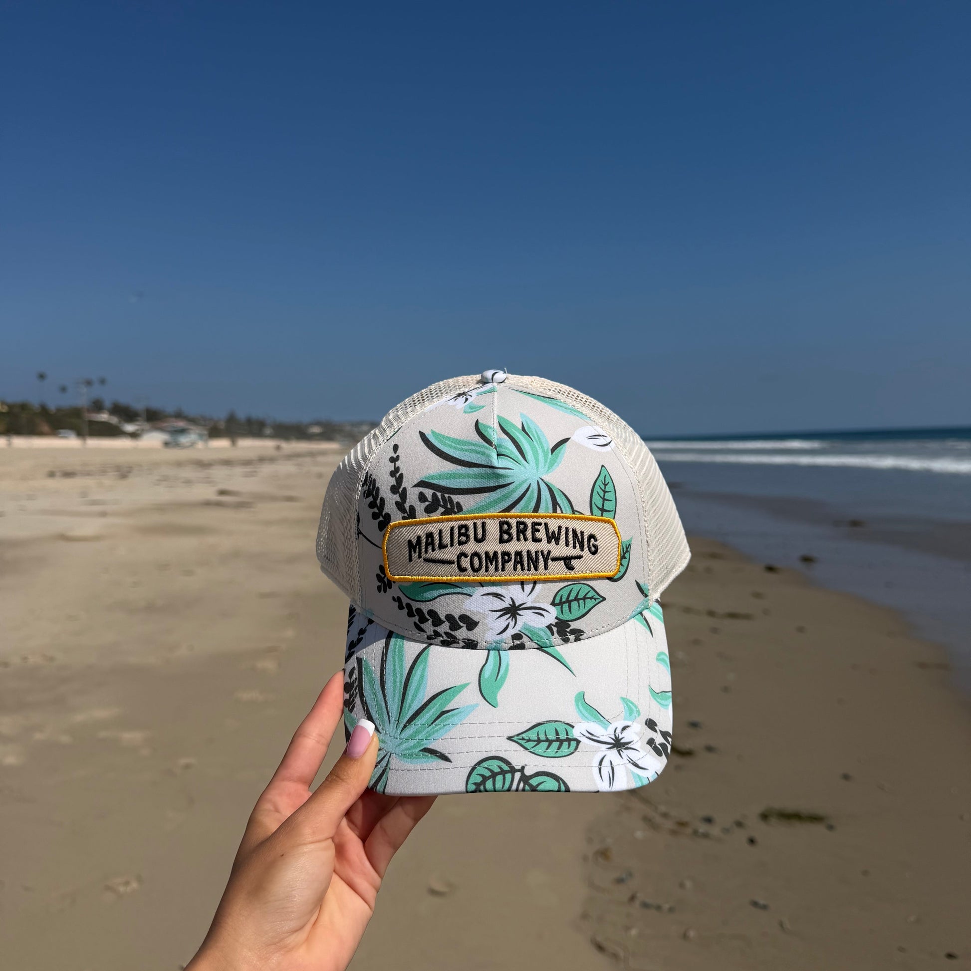 Cap with leaf pattern and 'Malibu Brewing Company' text held by a hand on a beach.