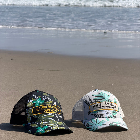 Two decorative hats with floral patterns with Malibu Brewing Company patches on a a sandy beach.