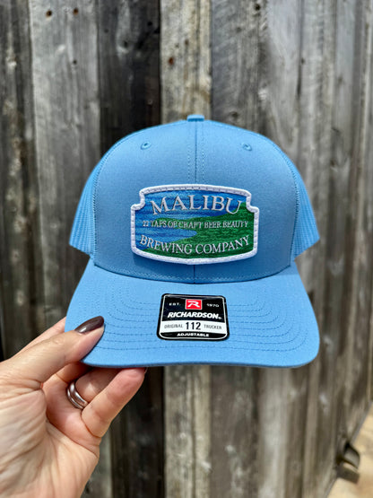 A hand holds a light blue Malibu City Sign Hat with an embroidered Malibu Brewing Company patch and a Richardson snap-back sticker on the brim, set against a wooden fence background.