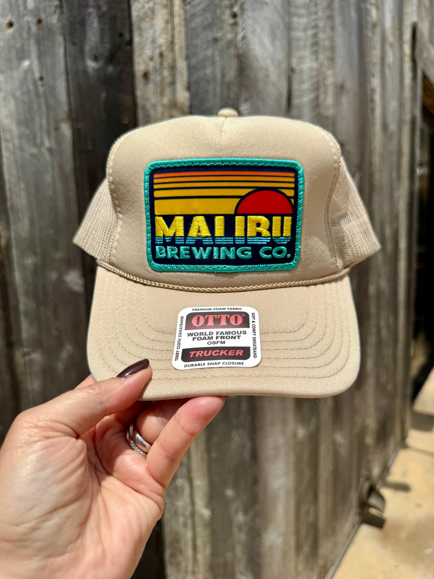 A hand holds the beige Otto Sunset Patch Hat with a vibrant Maui Brewing Co. patch. This trucker hat features mesh backing, a brim sticker, and is set against a wooden fence for a relaxed look.