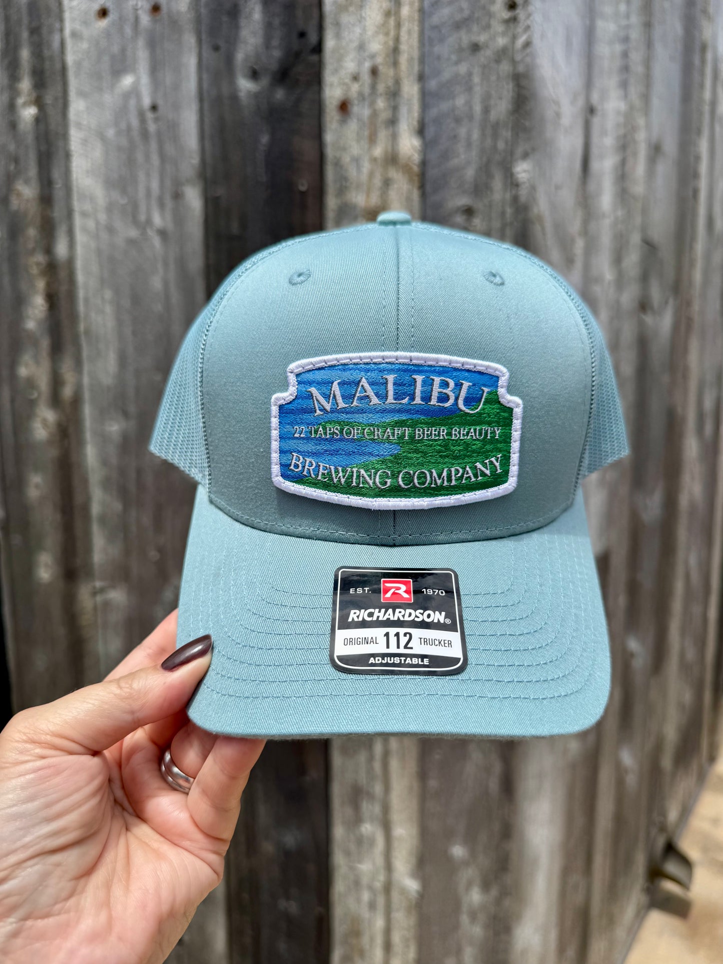 A hand holds a light blue Malibu City Sign Hat, a Richardson snapback featuring an embroidered Malibu Brewing Company patch with "27 Taps of Craft Beer Beauty," set against a weathered wood wall.