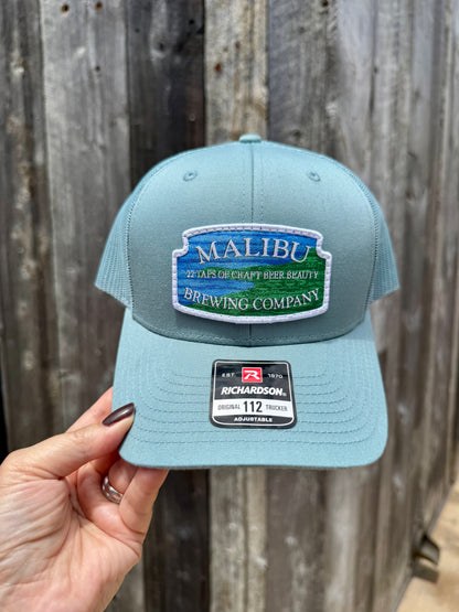 A hand holds a light blue Malibu City Sign Hat, a Richardson snapback featuring an embroidered Malibu Brewing Company patch with "27 Taps of Craft Beer Beauty," set against a weathered wood wall.