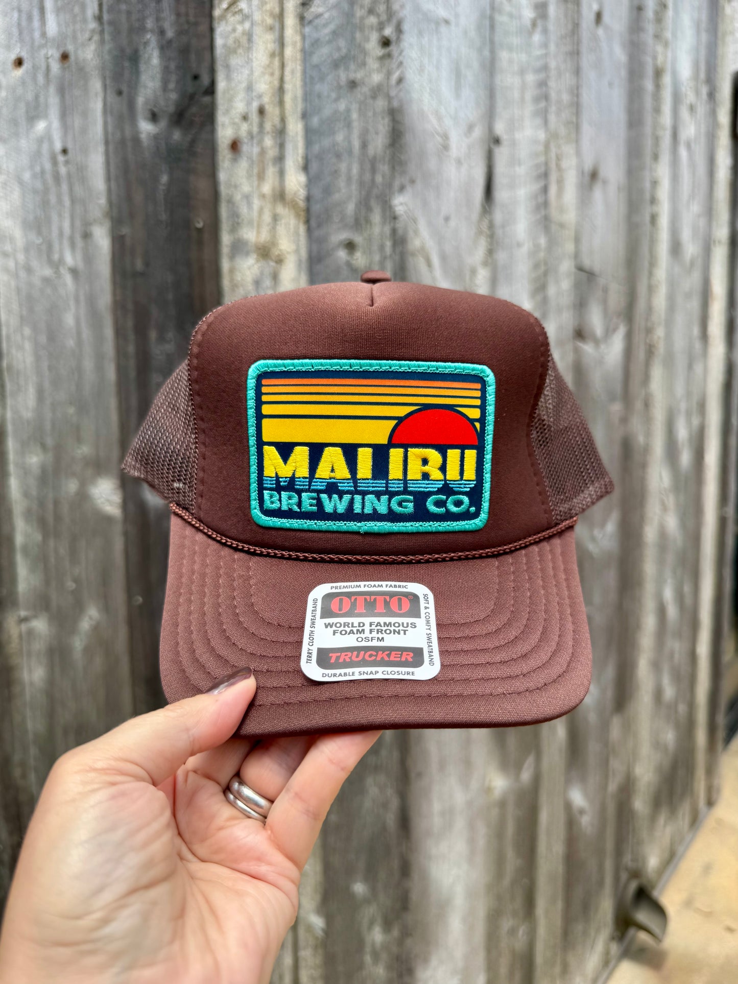 A hand holds the Otto Sunset Patch Hat, a brown mesh trucker hat featuring a vibrant Malibu Brewing Co. patch on the front, displayed against wooden planks.