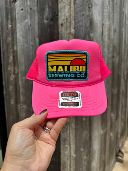 A hand holds the Otto Sunset Patch Hat, featuring a bright pink color, a vibrant Malibu Brewing Co. patch on the front, adjustable fit, and an Otto sticker on the brim, all set against a wooden background.