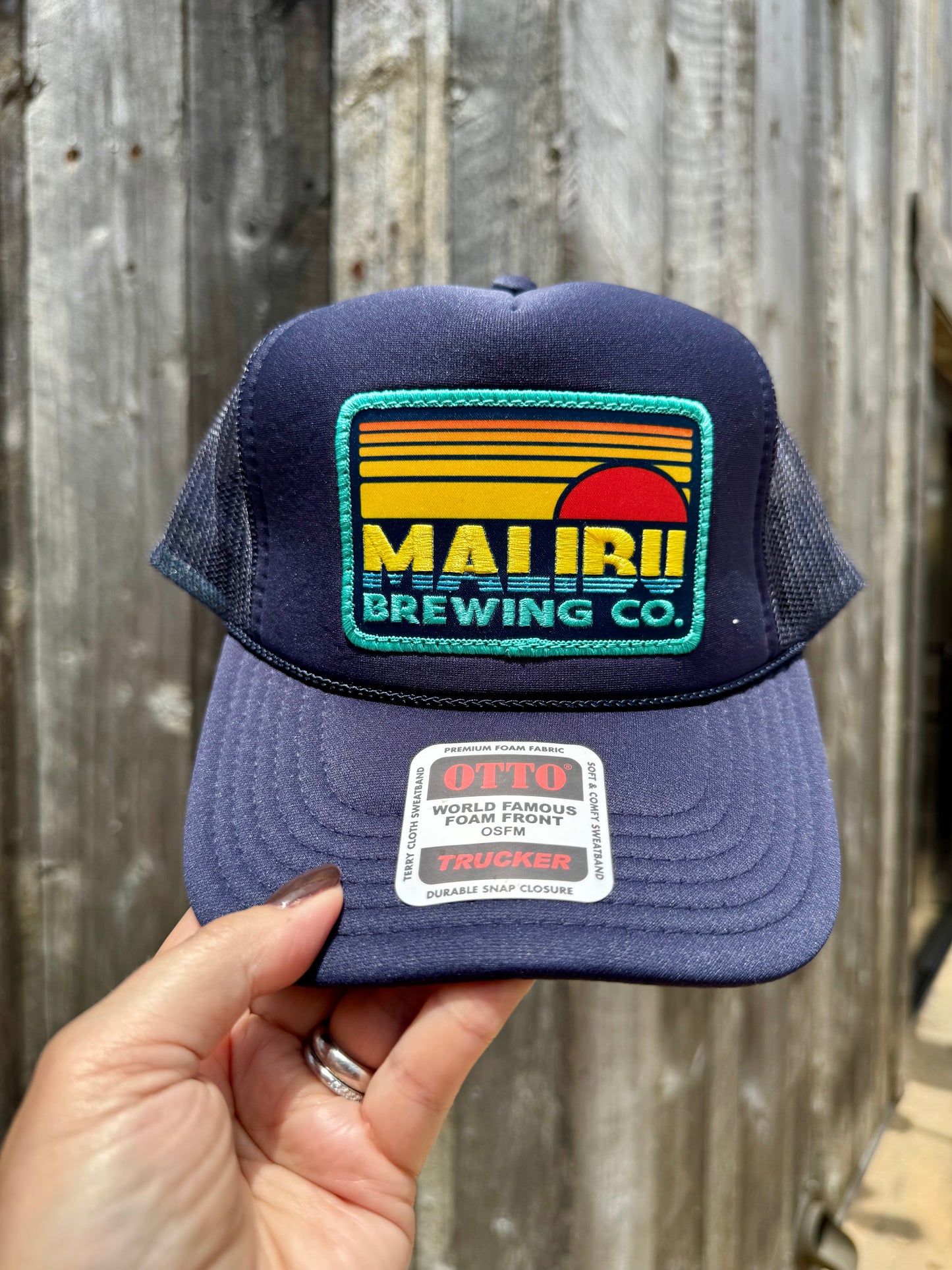 A hand holds the Otto Sunset Patch Hat in navy blue, featuring a colorful Maui Brewing Co. patch and OTTO sticker on the brim. This adjustable summer trucker hat is shown with a blurred wooden fence in the background.