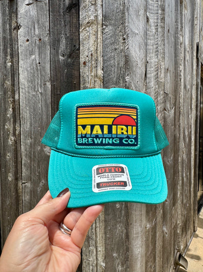 A hand holds the Otto Sunset Patch Hat, a summer cap with a vibrant MALIBU BREWING CO. patch and an OTTO WORLD FAMOUS TRUCKER sticker on the brim, set against a rustic wooden wall.