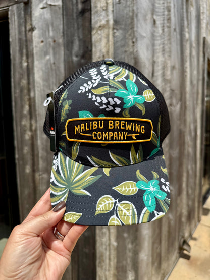A hand holds the Floral Trucker Hat, featuring black fabric with green and white tropical leaf patterns, an adjustable back, and a yellow "Malibu Brewing Company" patch, in front of a weathered wooden wall.