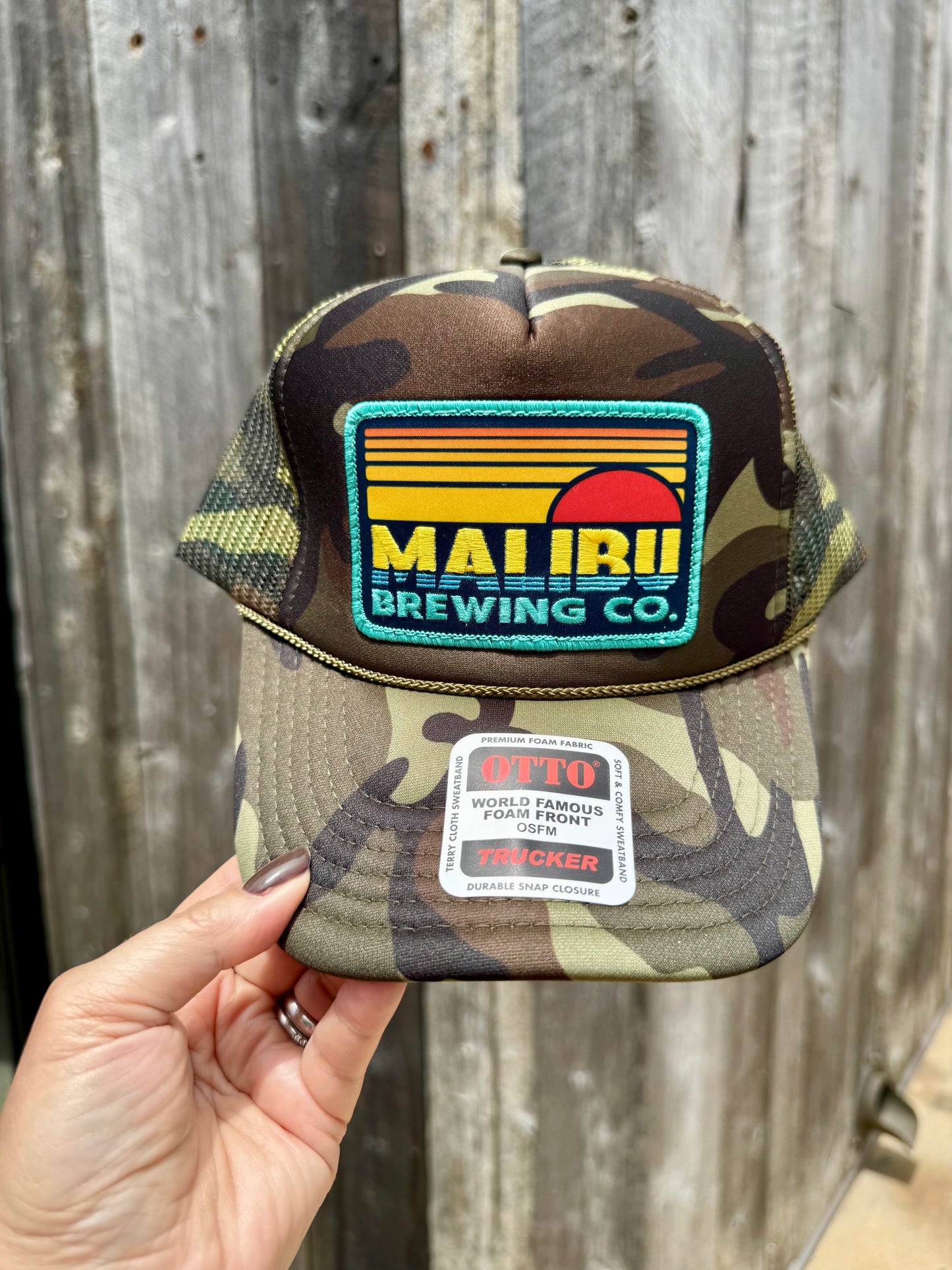 A hand holds the Otto Sunset Patch Hat, a camo summer trucker hat with mesh sides, an adjustable snap closure, and a colorful Maui Brewing Co. patch on the front; brim sticker reads OTTO Trucker.