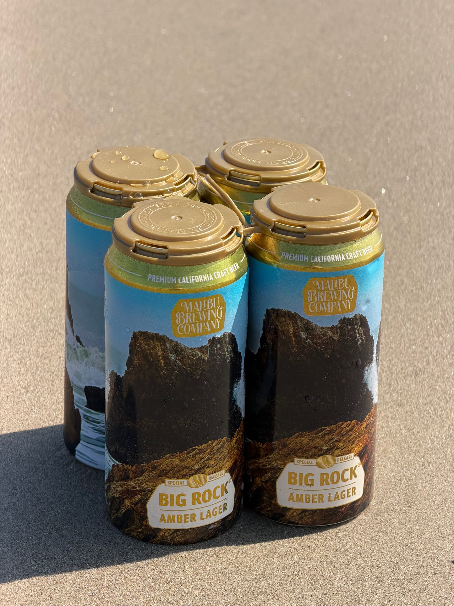 A Big Rock Amber Lager 4-Pack, a smooth Vienna Lager from Malibu Brewing Company, held together by tan plastic clips and resting on a sandy beach. The cans display scenic artwork of rocky ocean cliffs.