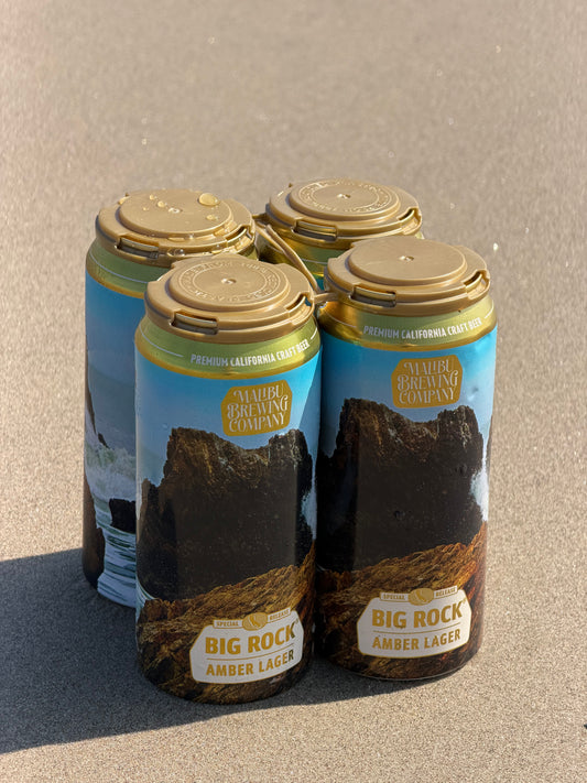 A Big Rock Amber Lager 4-Pack, a smooth Vienna Lager from Malibu Brewing Company, held together by tan plastic clips and resting on a sandy beach. The cans display scenic artwork of rocky ocean cliffs.