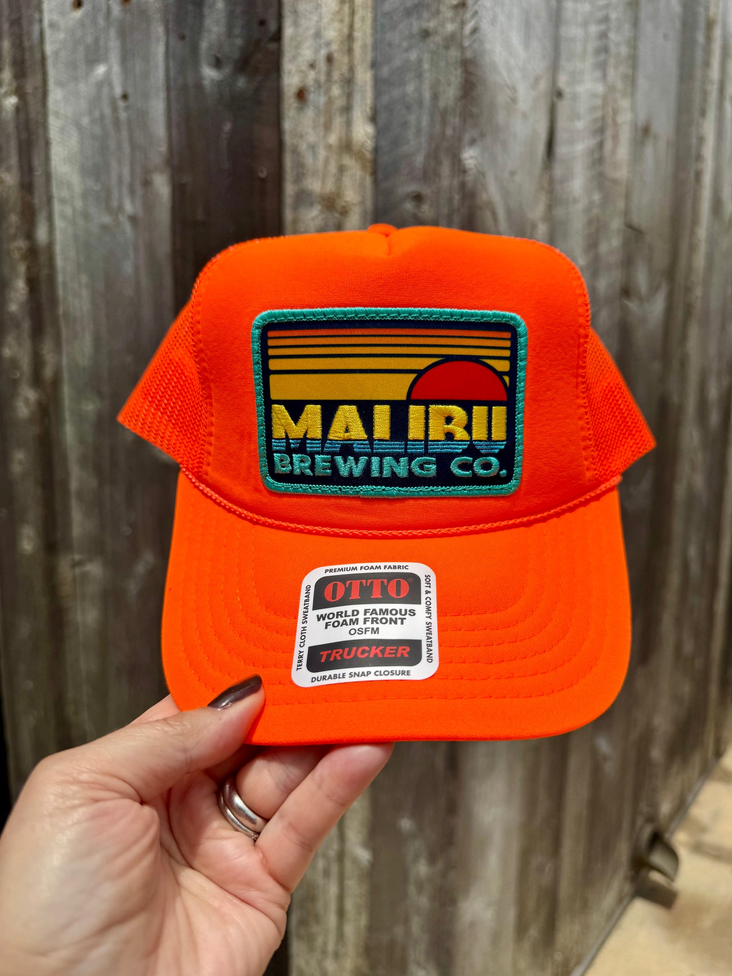 A hand holds the Otto Sunset Patch Hat, a vibrant orange summer trucker hat featuring a Maui Brewing Co. patch and an Otto World Famous Trucker sticker, with a weathered wooden fence in the background.