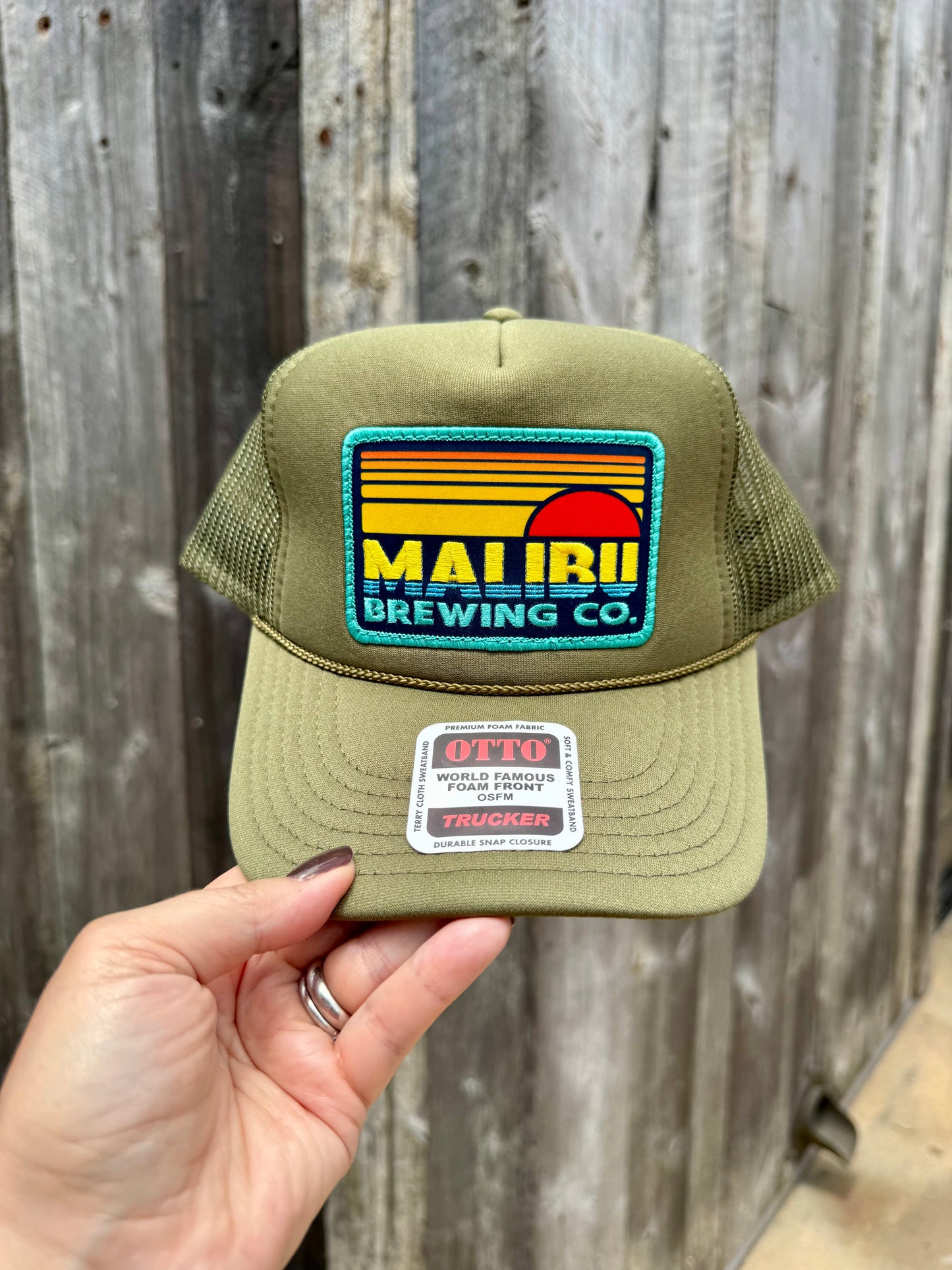 A hand holds the Otto Sunset Patch Hat in olive green, featuring a colorful Malibu Brewing Co. patch and an OTTO Trucker sticker on the brim, showing its adjustable fit against a wooden fence background.