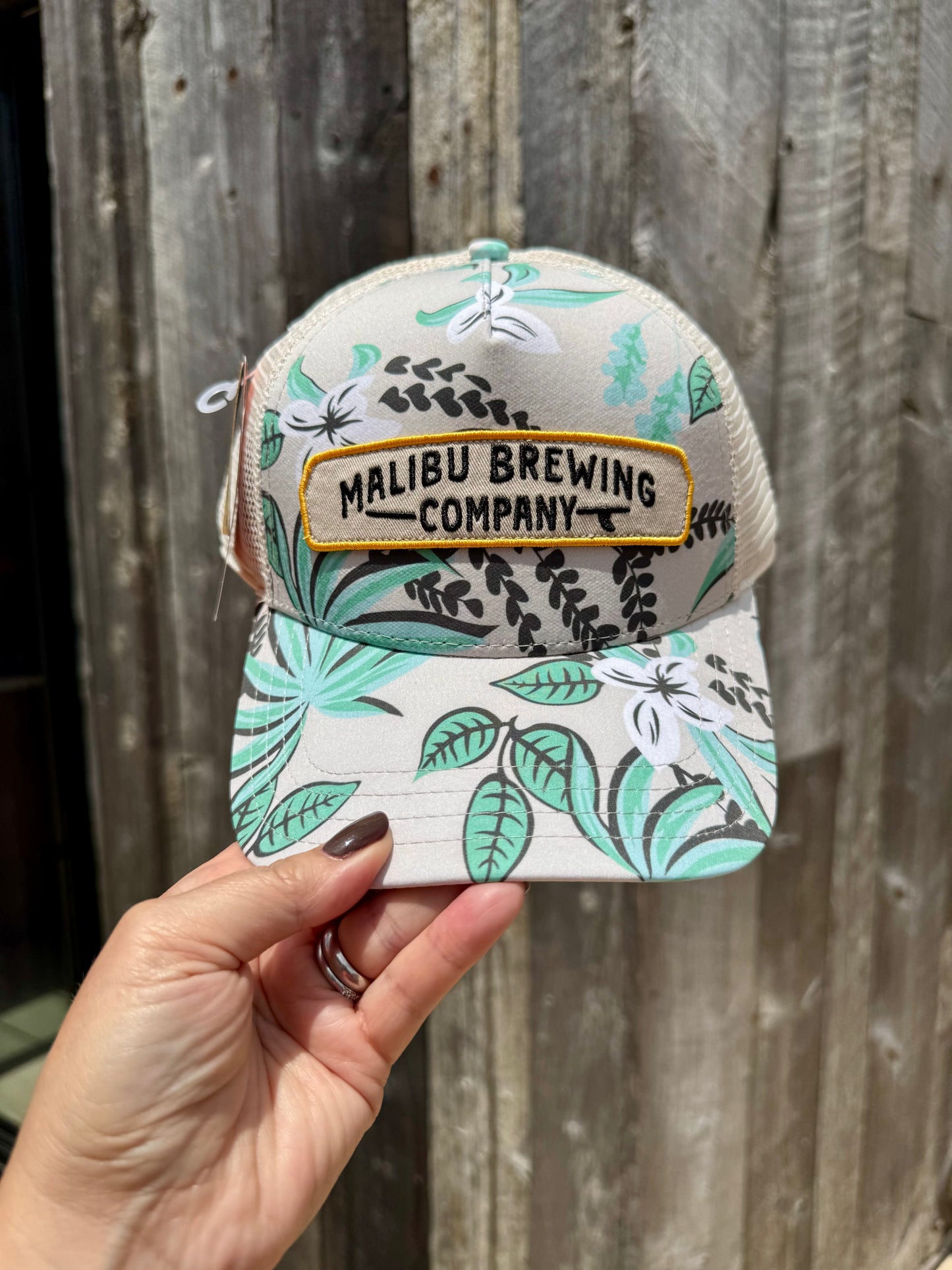 A hand holds the Floral Trucker Hat, featuring an embroidered Malibu Brewing Company patch, in front of a weathered wooden wall.