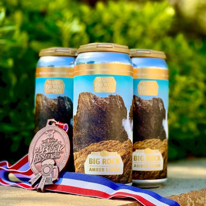 A 4-pack of Big Rock Amber Lager, an award-winning craft beer, is displayed outdoors with its bronze Missouri Beer Festival medal on a patriotic ribbon, set against a lively backdrop of blurred green foliage.
