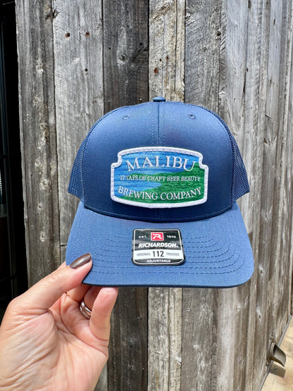 A hand holds a Malibu City Sign Hat with an embroidered "Malibu Brewing Company, 27 Miles of Craft Beer Beauty" patch. This Richardson snap-back has a black mesh back and brim sticker, set before a weathered wooden wall.