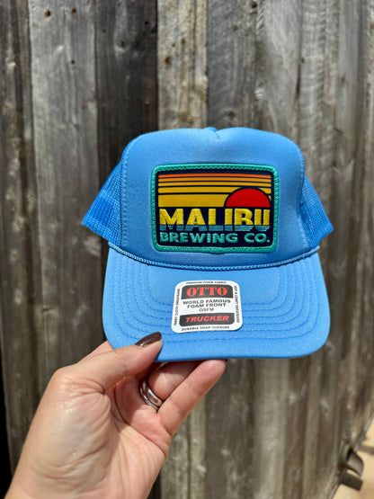 A hand holds the Otto Sunset Patch Hat, a blue summer cap featuring a Malibu Brewing Co. sunset logo, mesh back, and OTTO sticker, set against a wooden fence background.