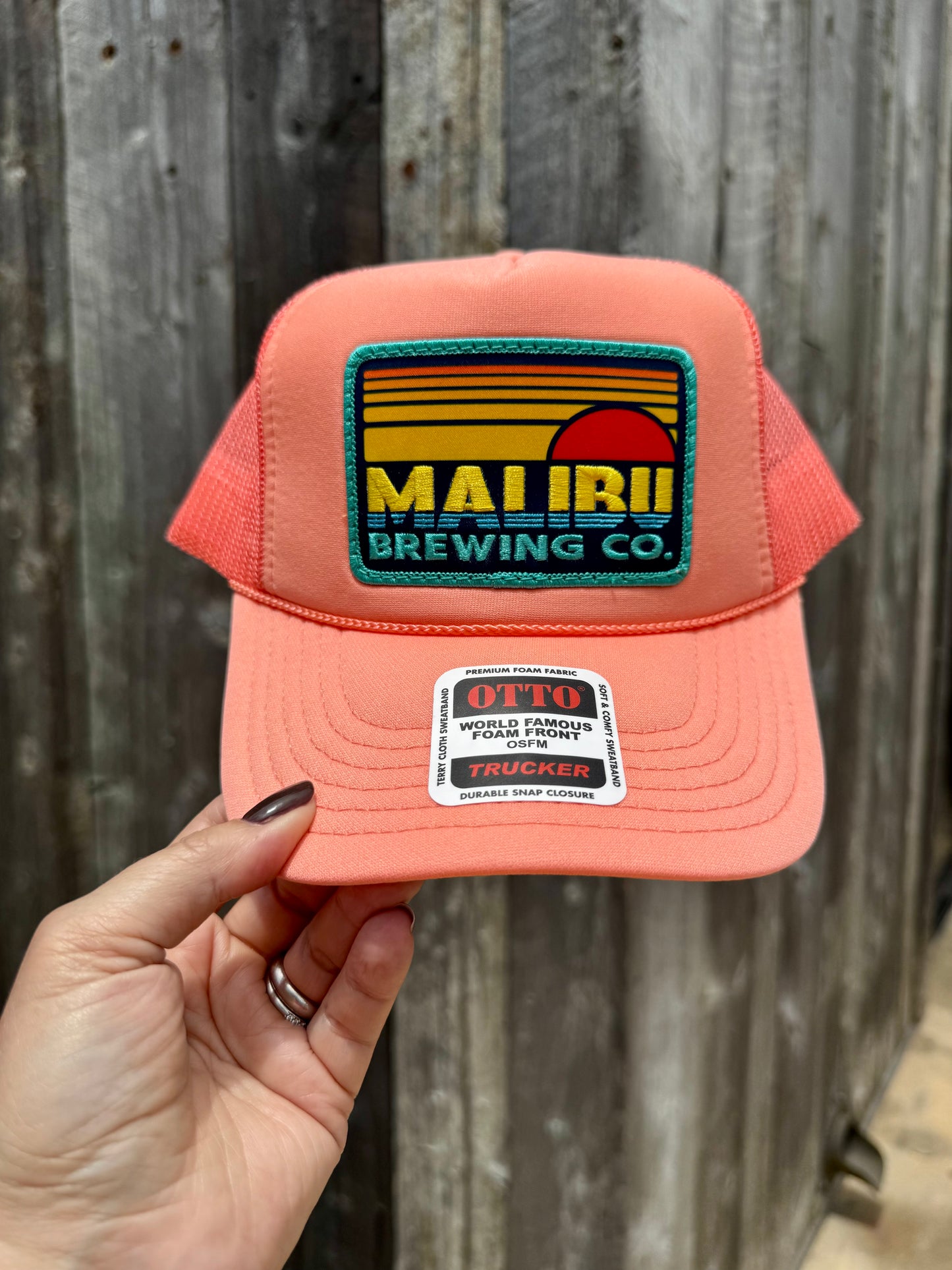 A hand holds the Otto Sunset Patch Hat, a coral classic trucker featuring a Malibu Brewing Co. sunset patch and an OTTO World Famous Pro Quality sticker on the front—an ideal summer accessory against wooden planks.