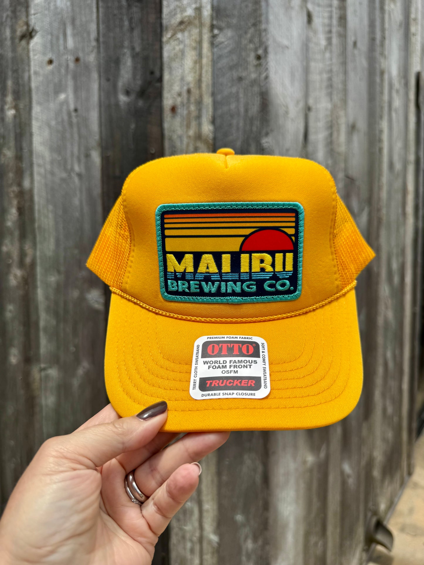 A hand holds the Otto Sunset Patch Hat, a bright yellow trucker cap with a colorful MAUI BREWING CO. patch and an OTTO World Famous Trucker sticker on the brim, set against a weathered wooden fence—your perfect summer accessory.