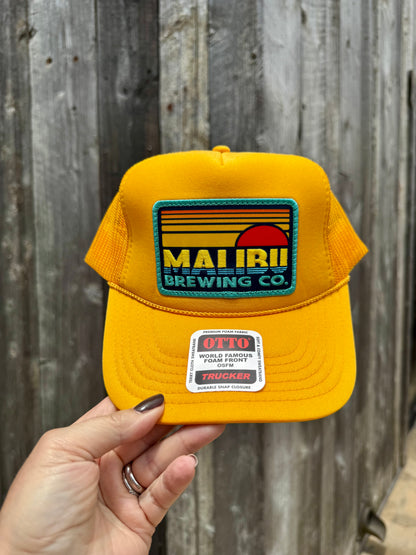 A hand holds the Otto Sunset Patch Hat, a bright yellow trucker cap with a colorful MAUI BREWING CO. patch and an OTTO World Famous Trucker sticker on the brim, set against a weathered wooden fence—your perfect summer accessory.