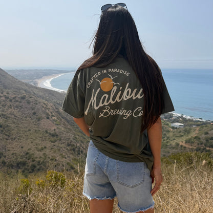 A woman with long dark hair, wearing the Crafted in Paradise Tee and denim shorts, stands on a sunny hillside overlooking the ocean and coastline.