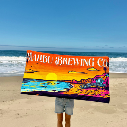 On a sandy California beach, someone holds the Beach Towel by Malibu Brewing Co. This colorful cotton terry towel displays "Malibu Brewing Co." with a sunset, ocean waves, birds, and a lifeguard hut among palm trees.
