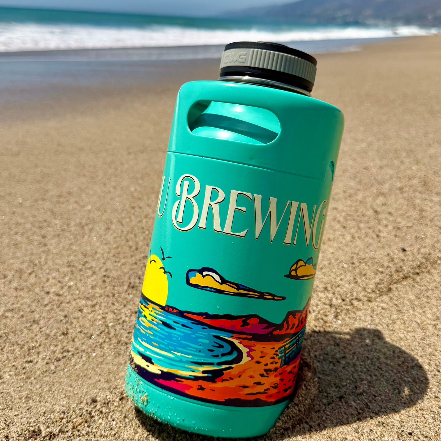 A Keg Growlers vacuum insulated growler in teal, featuring vibrant beach-themed artwork and the word BREWING, sits on a sandy beach with the ocean and mountains in the background.