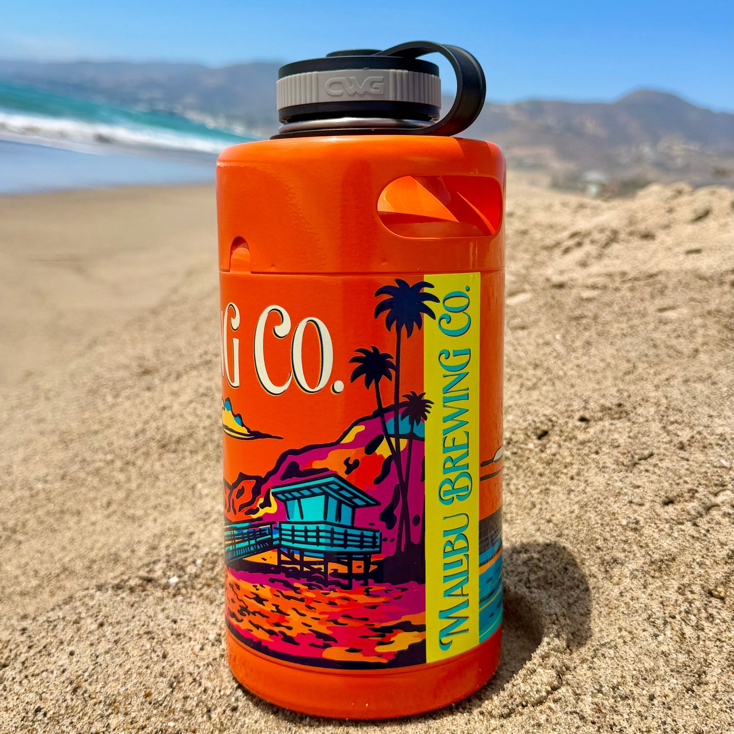 A vibrant orange Keg Growler from Malibu Brewing Co., featuring a beach hut and palm tree design, stands upright in the sand with the ocean and mountains in the background.