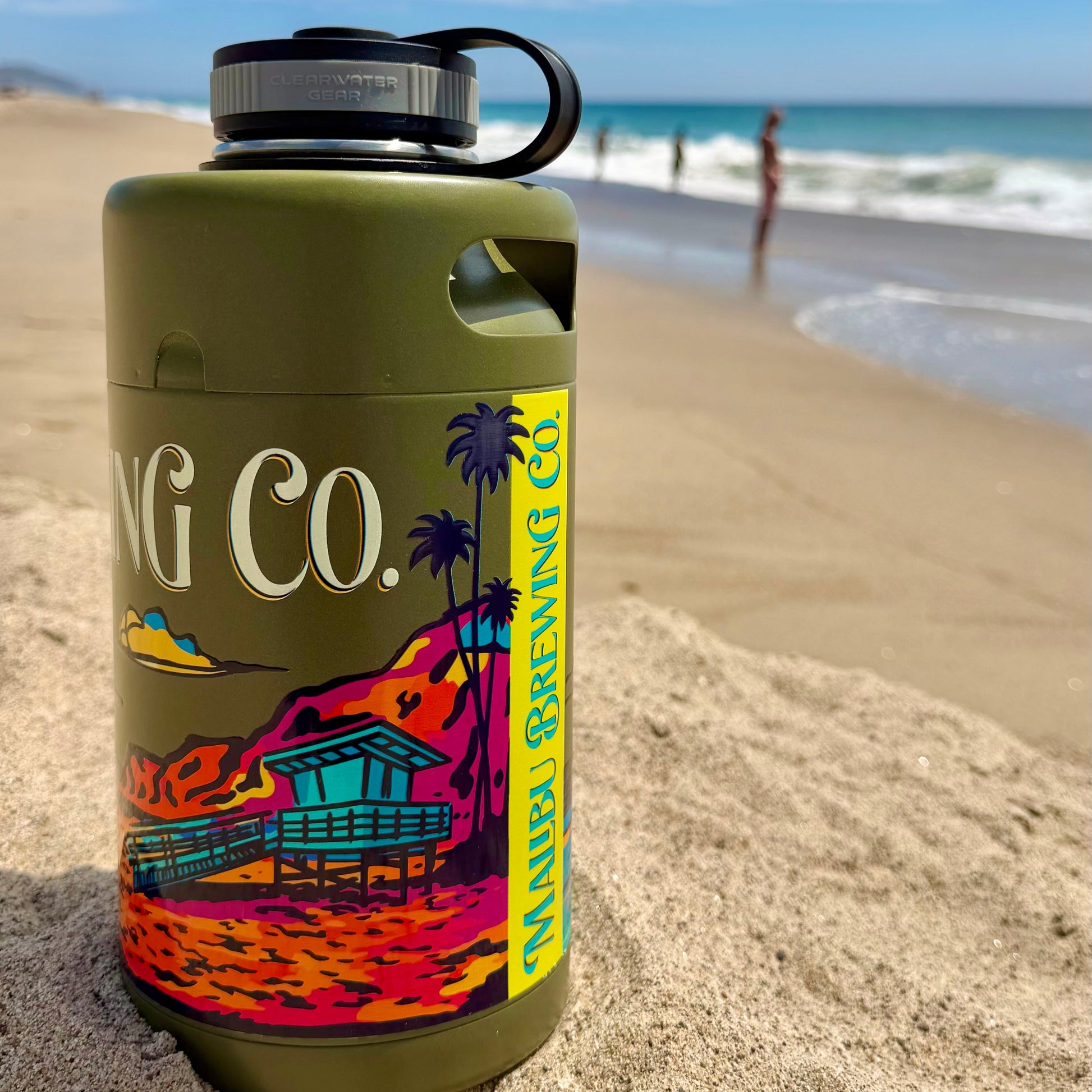 A Keg Growlers 64oz stainless water bottle from Malibu Brewing Co., featuring a green finish and a colorful beach design, sits on the sandy beach with waves and two people in the background.