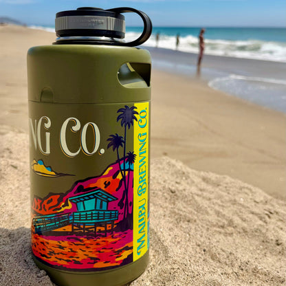 A Keg Growlers 64oz stainless water bottle from Malibu Brewing Co., featuring a green finish and a colorful beach design, sits on the sandy beach with waves and two people in the background.