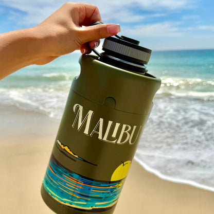 A hand holds a green Malibu Keg Growler decorated with a beach scene, with the ocean and sandy shore visible in the sunny background—perfect for your next adventure, this 64oz stainless Keg Growler keeps your drinks cold all day.
