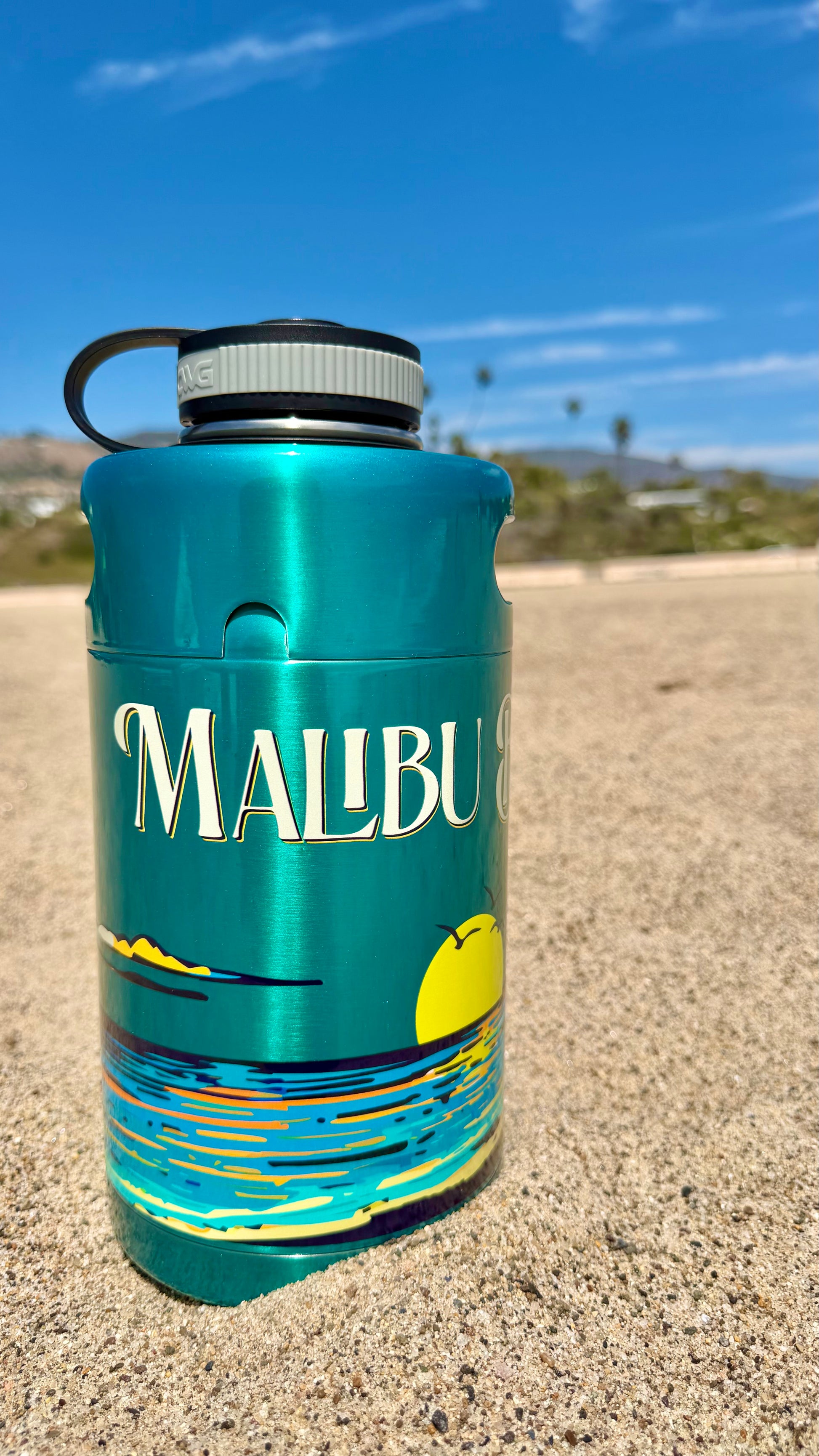 A Keg Growlers bottle in turquoise, featuring a Malibu beach sunset design, stands upright on sandy beach beneath a blue sky scattered with clouds and distant palm trees—its stylish 64oz stainless construction perfect for keeping your drinks cold during any coastal adventure.