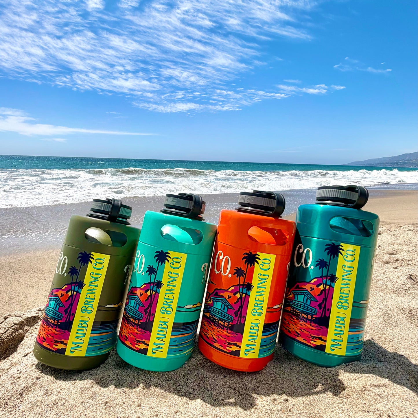 Four vibrant Keg Growlers from Malibu Brewing Co. stand upright on sandy beach, featuring beach-themed designs and bold colors, with a 64oz stainless growler included. Waves and a blue sky provide the perfect backdrop for these eye-catching bottles.