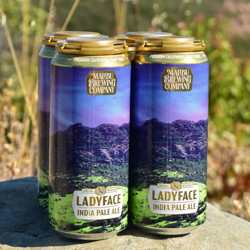 Four cans of Ladyface IPA rest on a rock outdoors, set against a blurred backdrop of green hills. The labels display a purple and green mountain scene, reflecting this West Coast IPA brewed with Simcoe and Mosaic hops.