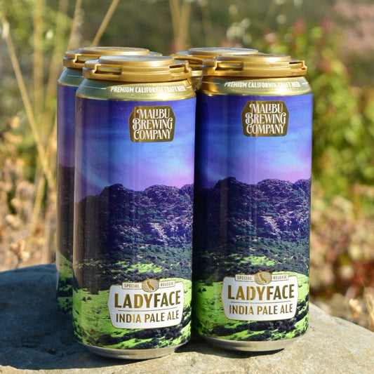 Four cans of Ladyface IPA rest on a rock outdoors, set against a blurred backdrop of green hills. The labels display a purple and green mountain scene, reflecting this West Coast IPA brewed with Simcoe and Mosaic hops.