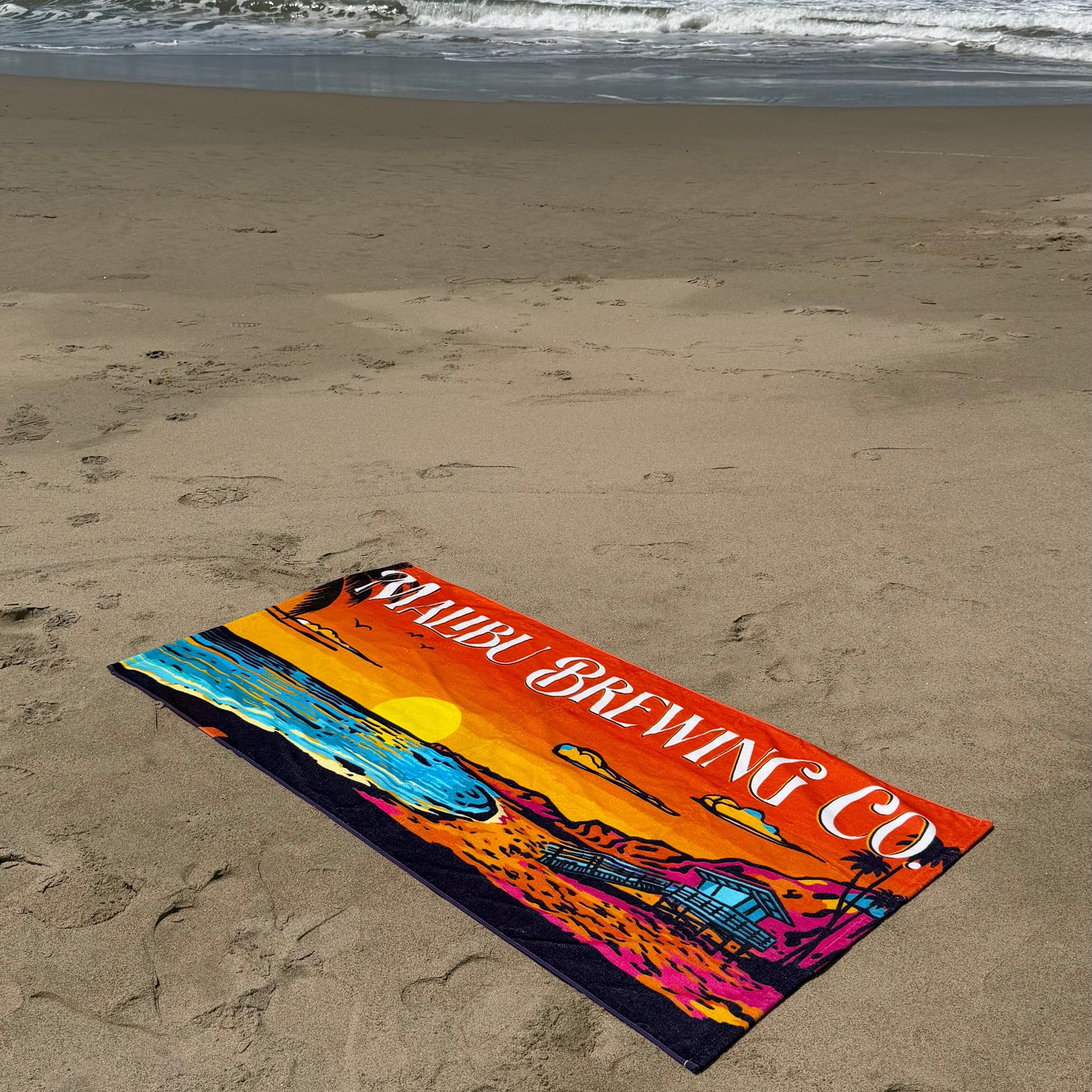 A colorful Beach Towel with sunset and waves design plus SAMUEL BREWING CO. text lies on sandy shores near the ocean—crafted from cotton terry, it’s perfect for relaxing by the water with gentle waves and footprints nearby.